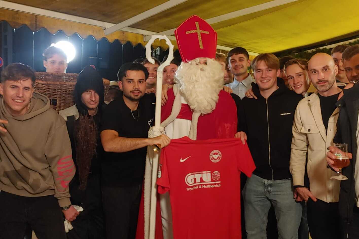 Gruppe junger Menschen posiert mit einem Santa Claus, der ein rotes T-Shirt h&auml;lt, in einer festlich dekorierten Umgebung.