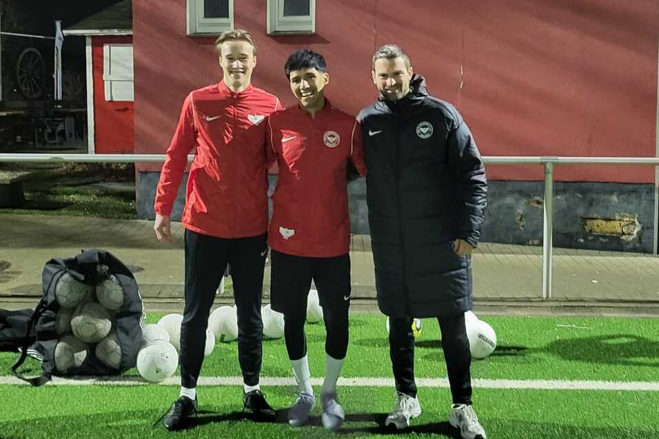 Drei Fu&szlig;ballspieler in Trainingskleidung stehen l&auml;chelnd auf einem Kunstrasenplatz vor einem Vereinsgeb&auml;ude bei Nacht.