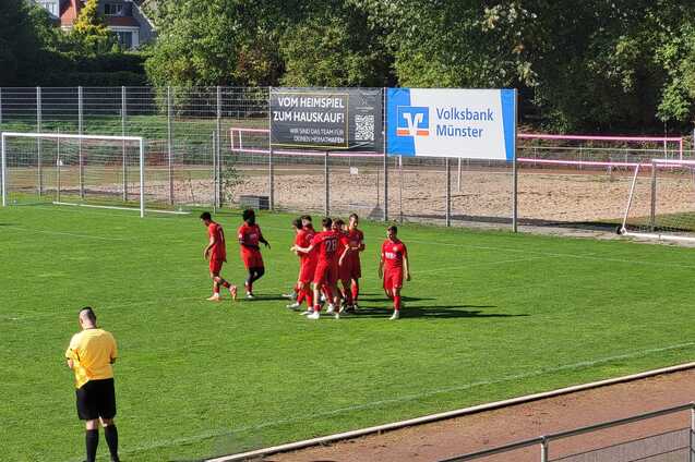 Spieler des Fu&szlig;ballteams in roten Jerseys feiern ein Tor auf einem gr&uuml;nen Sportplatz. Im Hintergrund stehen Tore und ein Werbeschild.