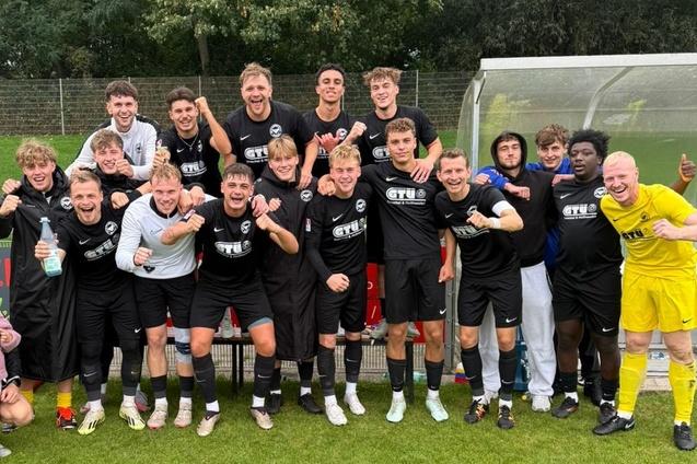 Gruppe von Fu&szlig;ballspielern in schwarzen Trikots steht jubelnd auf dem Spielfeld, einige tragen spezielle Trainingsbekleidung.
