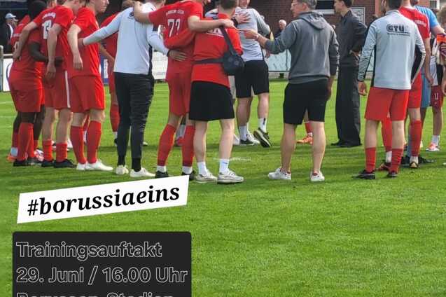 Gruppe von Spielern im roten Trikot versammelt auf einem Sportplatz, im Hintergrund ein Trainer und Banner mit Infos zum Training.