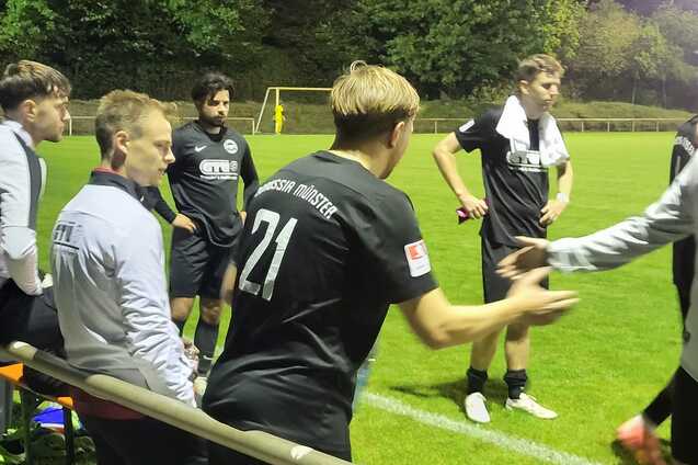 Eine Gruppe von Fu&szlig;ballspielern und Trainern sitzen an der Seitenlinie eines Fu&szlig;ballfeldes bei Nacht.