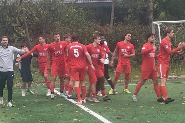 Gruppe von Fu&szlig;ballspielern in roten Trikots feiert auf dem gr&uuml;nen Sportplatz nach einem Spiel.
