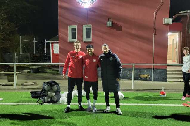 Drei Fu&szlig;ballspieler in Trainingskleidung stehen l&auml;chelnd auf einem Kunstrasenplatz vor einem Vereinsgeb&auml;ude bei Nacht.