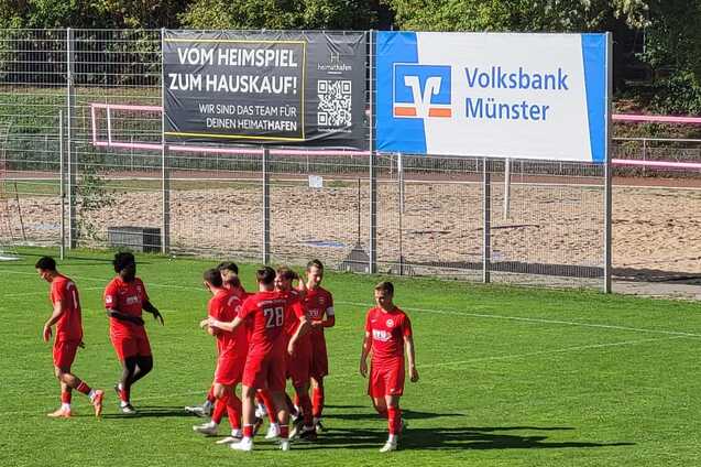 Spieler des Fu&szlig;ballteams in roten Jerseys feiern ein Tor auf einem gr&uuml;nen Sportplatz. Im Hintergrund stehen Tore und ein Werbeschild.