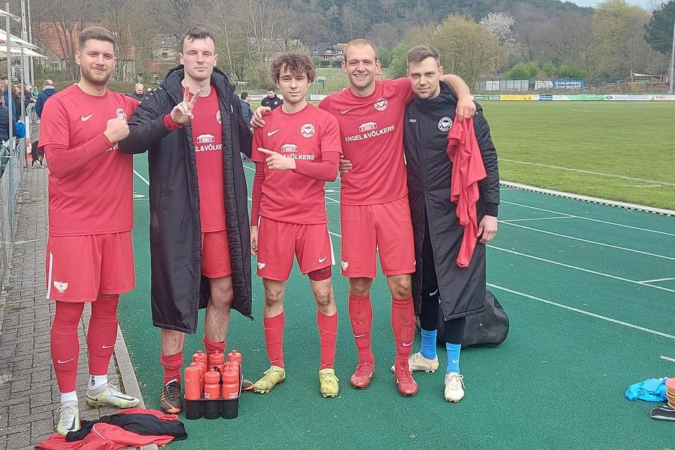 F&uuml;nf Fu&szlig;ballspieler in roten Trikots posieren l&auml;chelnd nach einem Spiel neben einem Sportplatz.