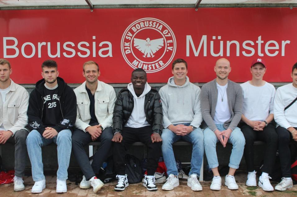 Acht M&auml;nner sitzen auf einer Bank am Spielfeldrand, hinter ihnen ist ein rotes Banner mit &bdquo;Borussia M&uuml;nster&ldquo;.