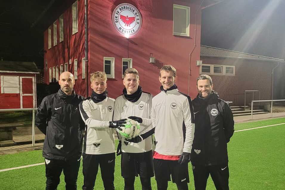 Gruppierung von f&uuml;nf Fu&szlig;ballspielern in Trainingskleidung auf einem beleuchteten Kunstrasenplatz vor einem Vereinsgeb&auml;ude.
