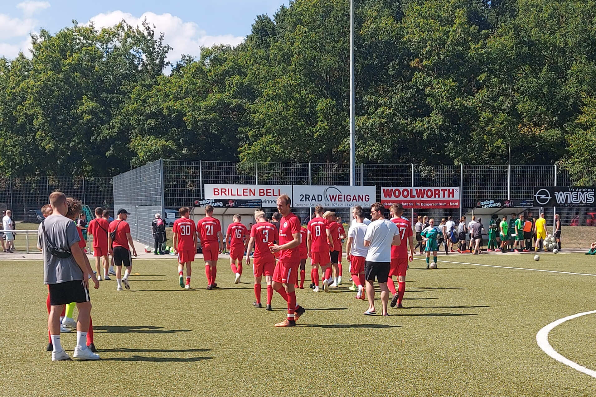 Gruppe von Fu&szlig;ballspielern in roten Trikots geht &uuml;ber einen Kunstrasenplatz, umgeben von B&auml;umen und Zuschauerb&auml;nken.