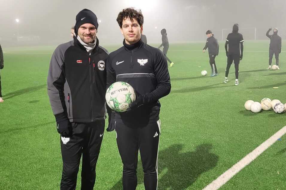 Zwei Fu&szlig;ballspieler posieren mit einem Ball auf einem Trainingsplatz bei nebligem Wetter, andere Spieler im Hintergrund.
