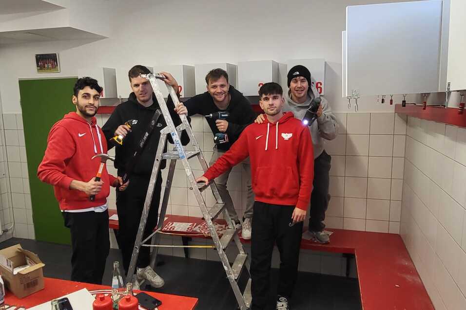 F&uuml;nf Personen in roten Sweatshirts posieren auf einer Bank neben einer Leiter in einem Umkleideraum.