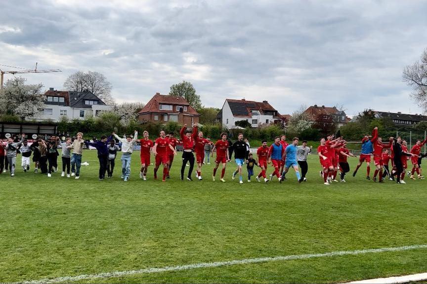 Gruppierung von Spielern und Fans, die gemeinsam auf einem Fu&szlig;ballplatz jubeln und tanzen, umgeben von Wohnh&auml;usern.