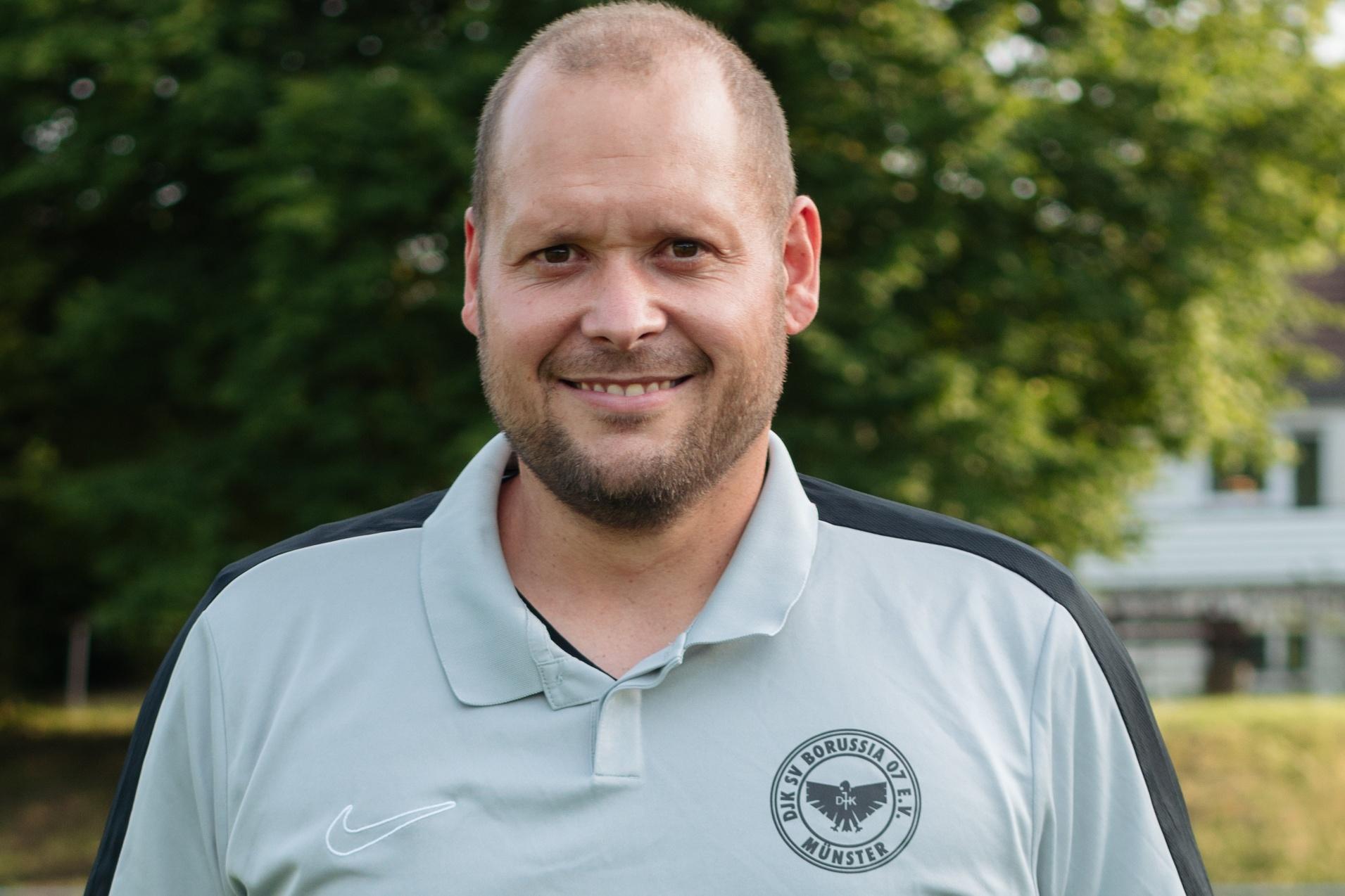 L&auml;chelnder Trainer in hellgrauem Polo-Shirt vor einem Sportplatz mit B&auml;umen im Hintergrund.