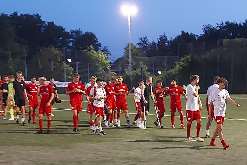 Gruppe von Fu&szlig;ballspielern in roten und blauen Trikots auf einem Kunstrasenplatz bei D&auml;mmerung.