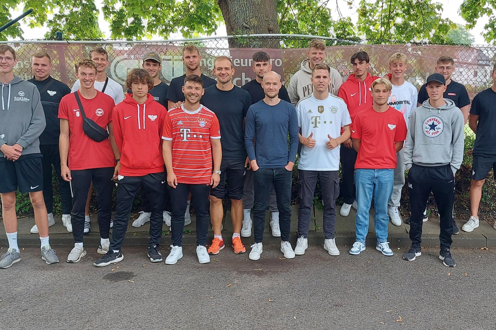 Gruppe von 20 jungen M&auml;nnern in Trainingsanz&uuml;gen steht auf einem Platz vor einem Zaun mit Werbebanner.