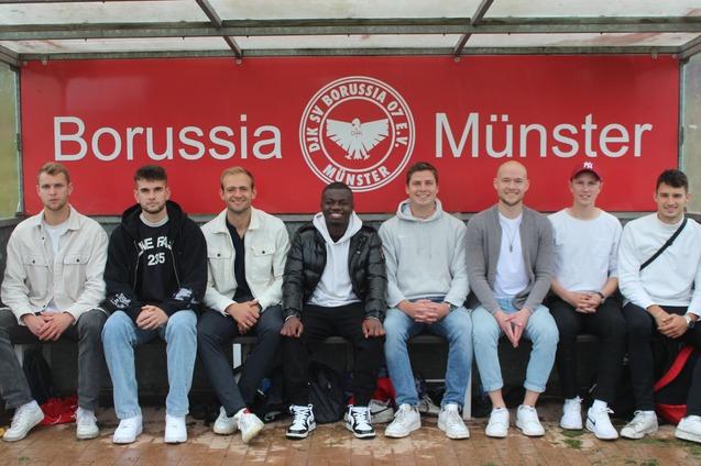 Acht M&auml;nner sitzen auf einer Bank am Spielfeldrand, hinter ihnen ist ein rotes Banner mit &bdquo;Borussia M&uuml;nster&ldquo;.