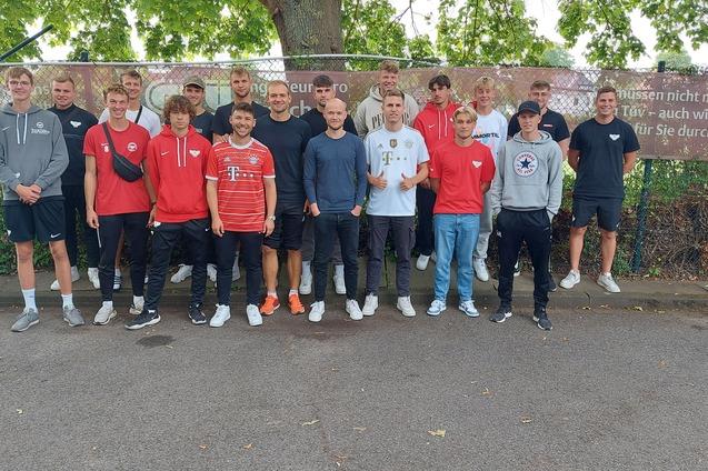 Gruppe von 20 jungen M&auml;nnern in Trainingsanz&uuml;gen steht auf einem Platz vor einem Zaun mit Werbebanner.