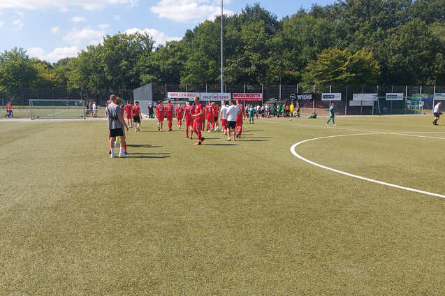 Gruppe von Fu&szlig;ballspielern in roten Trikots geht &uuml;ber einen Kunstrasenplatz, umgeben von B&auml;umen und Zuschauerb&auml;nken.