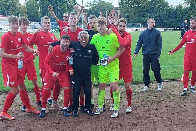 Gruppe jubelnder Fu&szlig;ballspieler in roten Trikots, umrahmt von einem Torwart in gr&uuml;ner Ausr&uuml;stung auf dem Spielfeld.