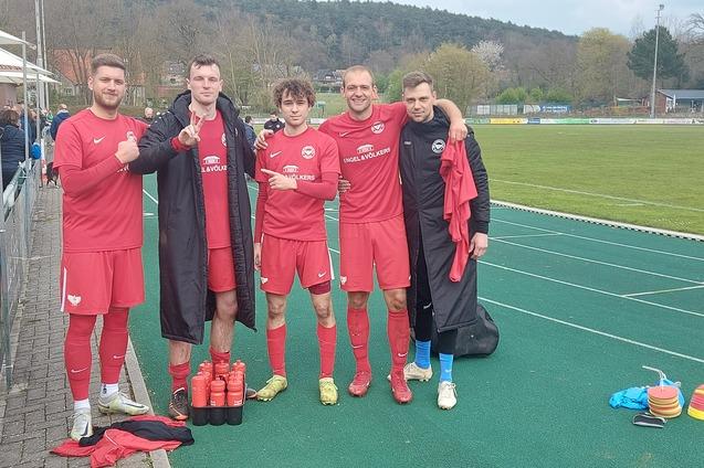 F&uuml;nf Fu&szlig;ballspieler in roten Trikots posieren l&auml;chelnd nach einem Spiel neben einem Sportplatz.