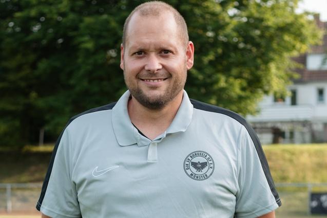 L&auml;chelnder Trainer in hellgrauem Polo-Shirt vor einem Sportplatz mit B&auml;umen im Hintergrund.