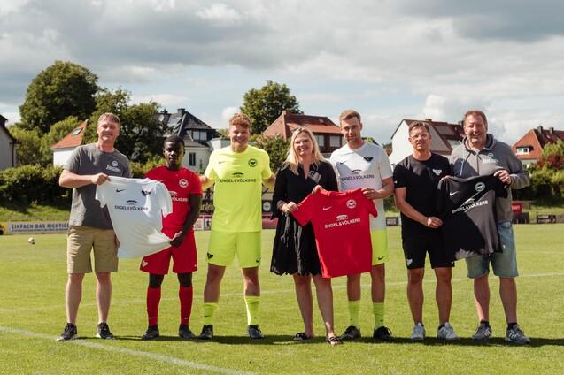 Sechs Personen auf einem Fu&szlig;ballfeld halten Trikots in verschiedenen Farben, im Hintergrund ist eine Wohngegend sichtbar.