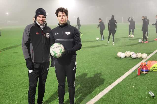 Zwei Fu&szlig;ballspieler posieren mit einem Ball auf einem Trainingsplatz bei nebligem Wetter, andere Spieler im Hintergrund.