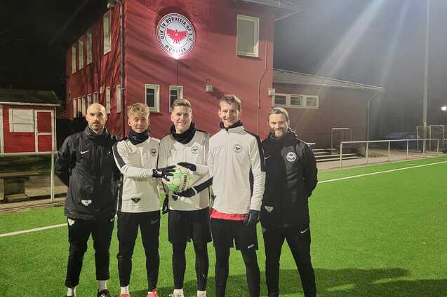 Gruppierung von f&uuml;nf Fu&szlig;ballspielern in Trainingskleidung auf einem beleuchteten Kunstrasenplatz vor einem Vereinsgeb&auml;ude.