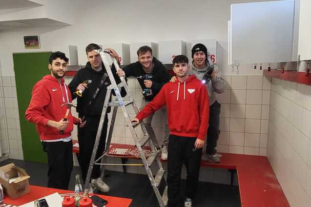 F&uuml;nf Personen in roten Sweatshirts posieren auf einer Bank neben einer Leiter in einem Umkleideraum.