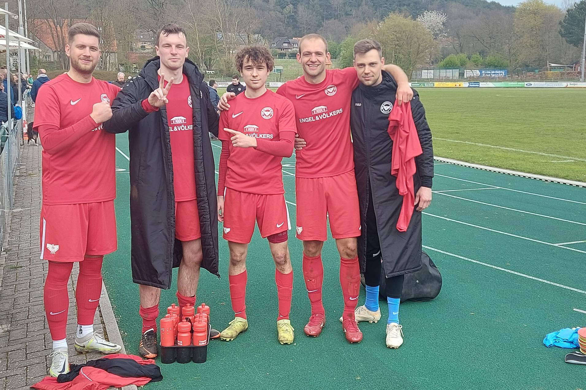 F&uuml;nf Fu&szlig;ballspieler in roten Trikots posieren l&auml;chelnd nach einem Spiel neben einem Sportplatz.
