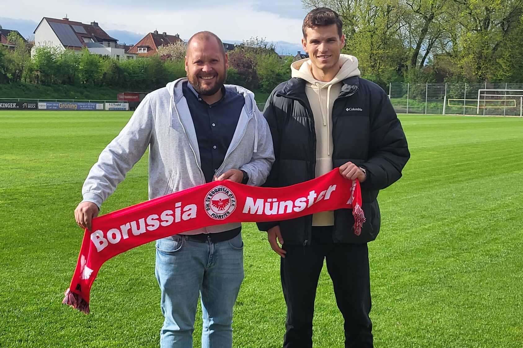 Zwei M&auml;nner stehen auf einem Fu&szlig;ballplatz und halten einen roten Schal mit der Aufschrift "Borussia M&uuml;nster".