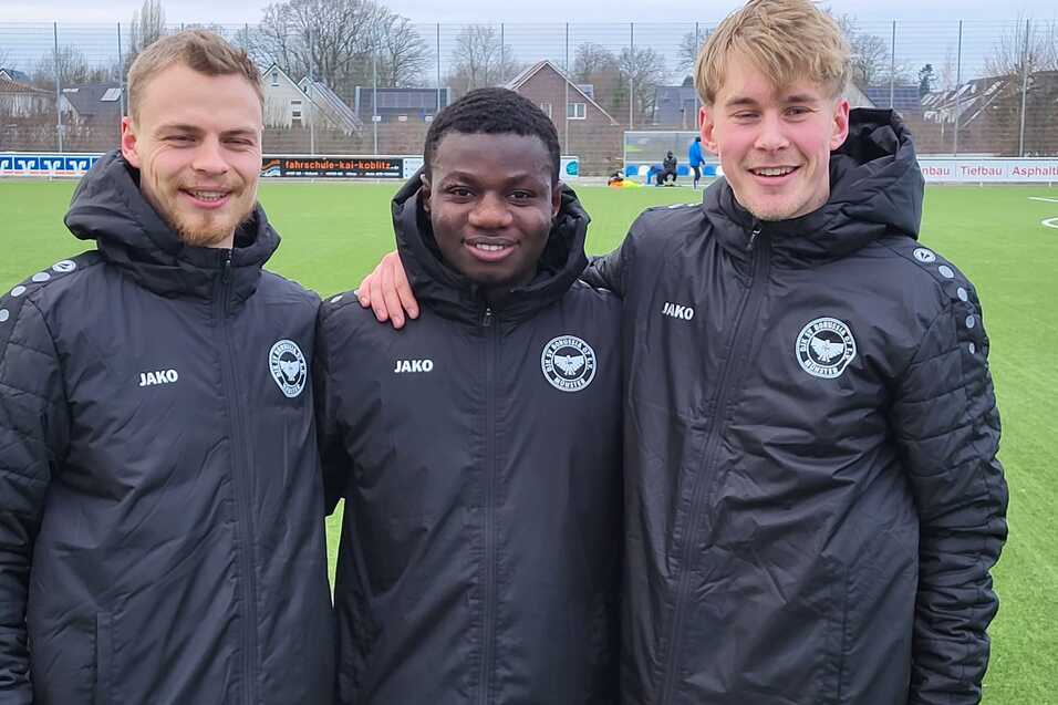 Drei Fu&szlig;ballspieler in schwarzen Trainingsjacken stehen l&auml;chelnd auf einem Rasenplatz.