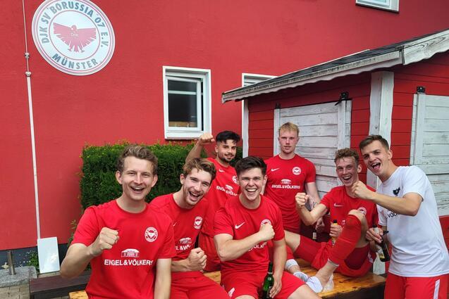 Sechs Fu&szlig;ballspieler in roten Trikots posieren l&auml;chelnd auf einer Bank vor einem roten Vereinsgeb&auml;ude, mit Getr&auml;nken in der Hand.