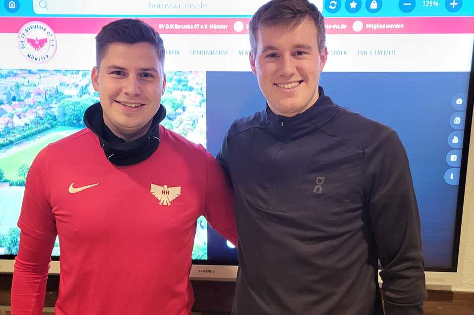 Zwei junge M&auml;nner stehen nebeneinander vor einem Bildschirm mit einer Stadtansicht, einer tr&auml;gt ein rotes Shirt, der andere ein graues.