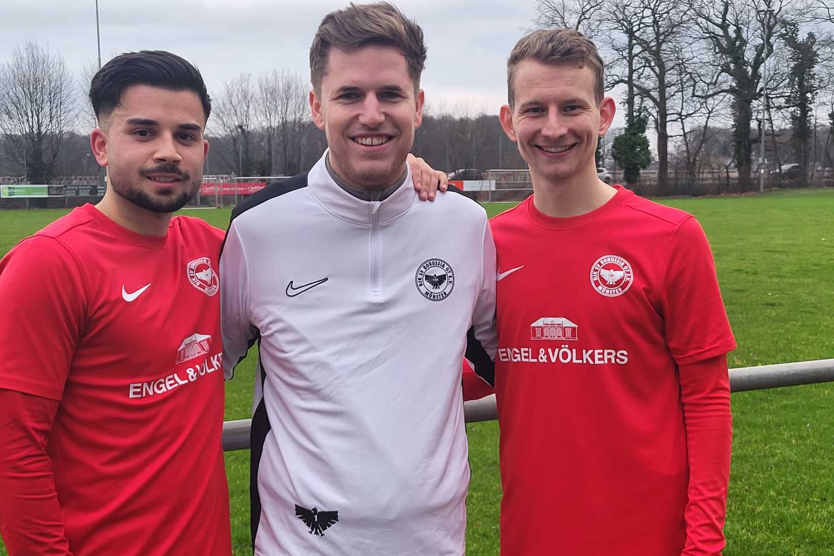Drei junge M&auml;nner in roten Fu&szlig;balltrikots stehen l&auml;chelnd auf einem Sportplatz mit gr&uuml;ner Wiese im Hintergrund.