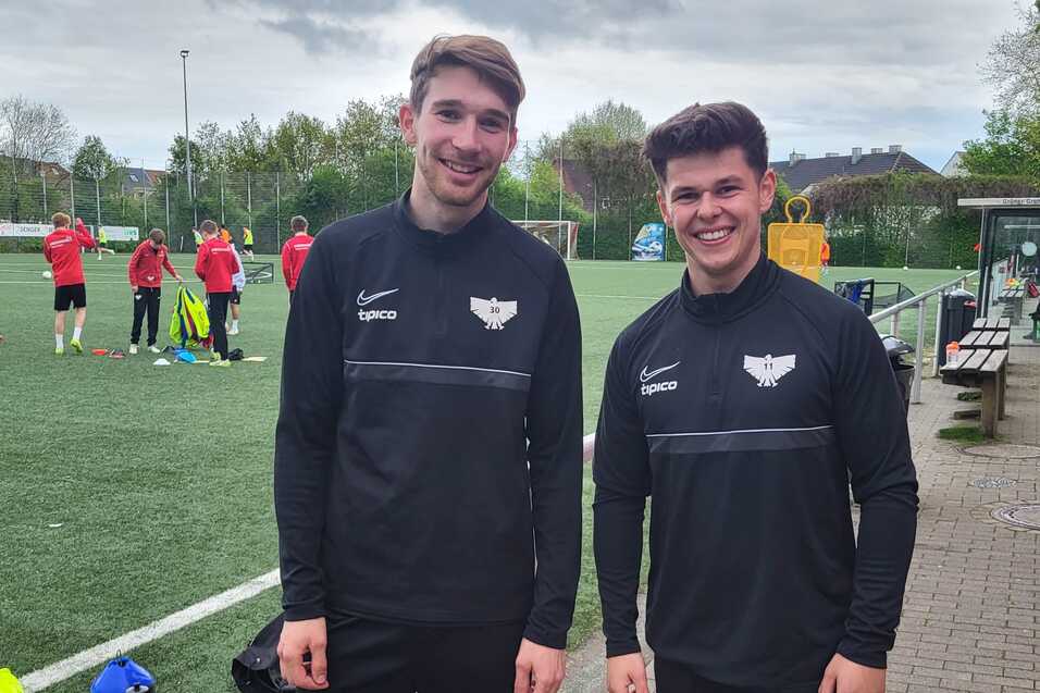Zwei junge M&auml;nner in Sportkleidung stehen l&auml;chelnd auf einem Fu&szlig;ballplatz, im Hintergrund sind weitere Spieler aktiv.