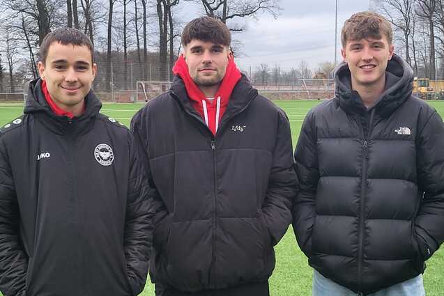 Drei junge M&auml;nner stehen auf einem Fu&szlig;ballplatz, umgeben von B&auml;umen, mit Sportjacken und verschiedenen Outfits.