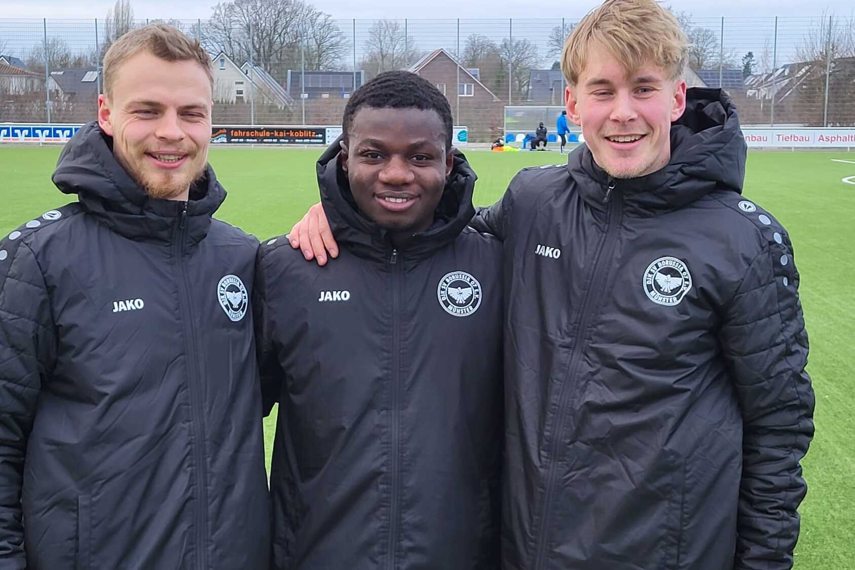 Drei Fu&szlig;ballspieler in schwarzen Trainingsjacken stehen l&auml;chelnd auf einem Rasenplatz.