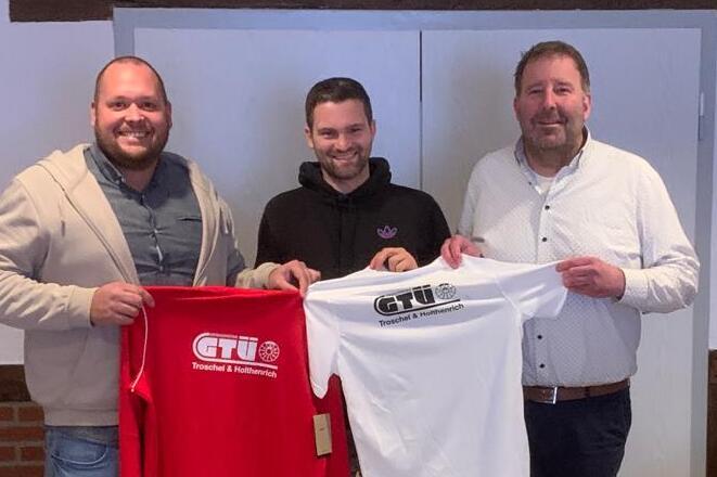 Drei M&auml;nner stehen vor einer geschlossenen T&uuml;r und halten rote und wei&szlig;e T-Shirts mit dem Logo &bdquo;GT&Uuml;&ldquo; in der Hand.