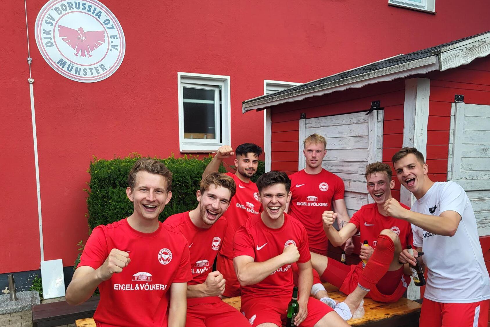 Sechs Fu&szlig;ballspieler in roten Trikots posieren l&auml;chelnd auf einer Bank vor einem roten Vereinsgeb&auml;ude, mit Getr&auml;nken in der Hand.