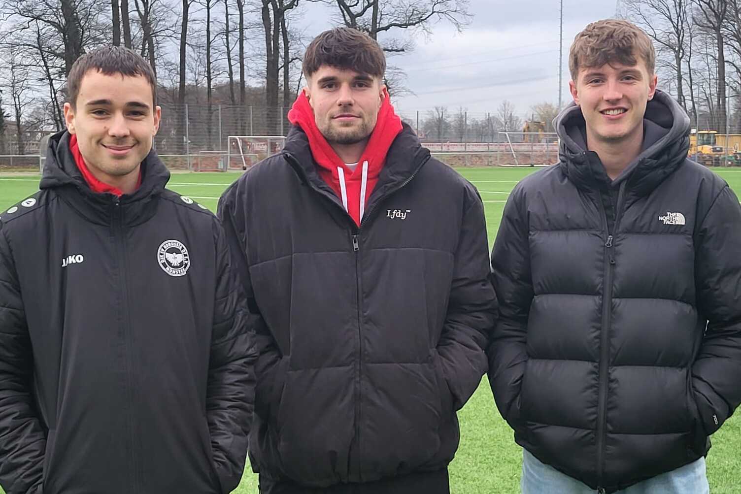 Drei junge M&auml;nner stehen auf einem Fu&szlig;ballplatz, umgeben von B&auml;umen, mit Sportjacken und verschiedenen Outfits.