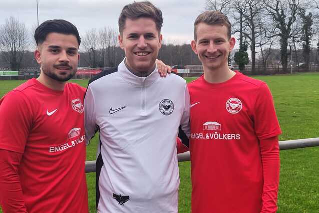 Drei junge M&auml;nner in roten Fu&szlig;balltrikots stehen l&auml;chelnd auf einem Sportplatz mit gr&uuml;ner Wiese im Hintergrund.