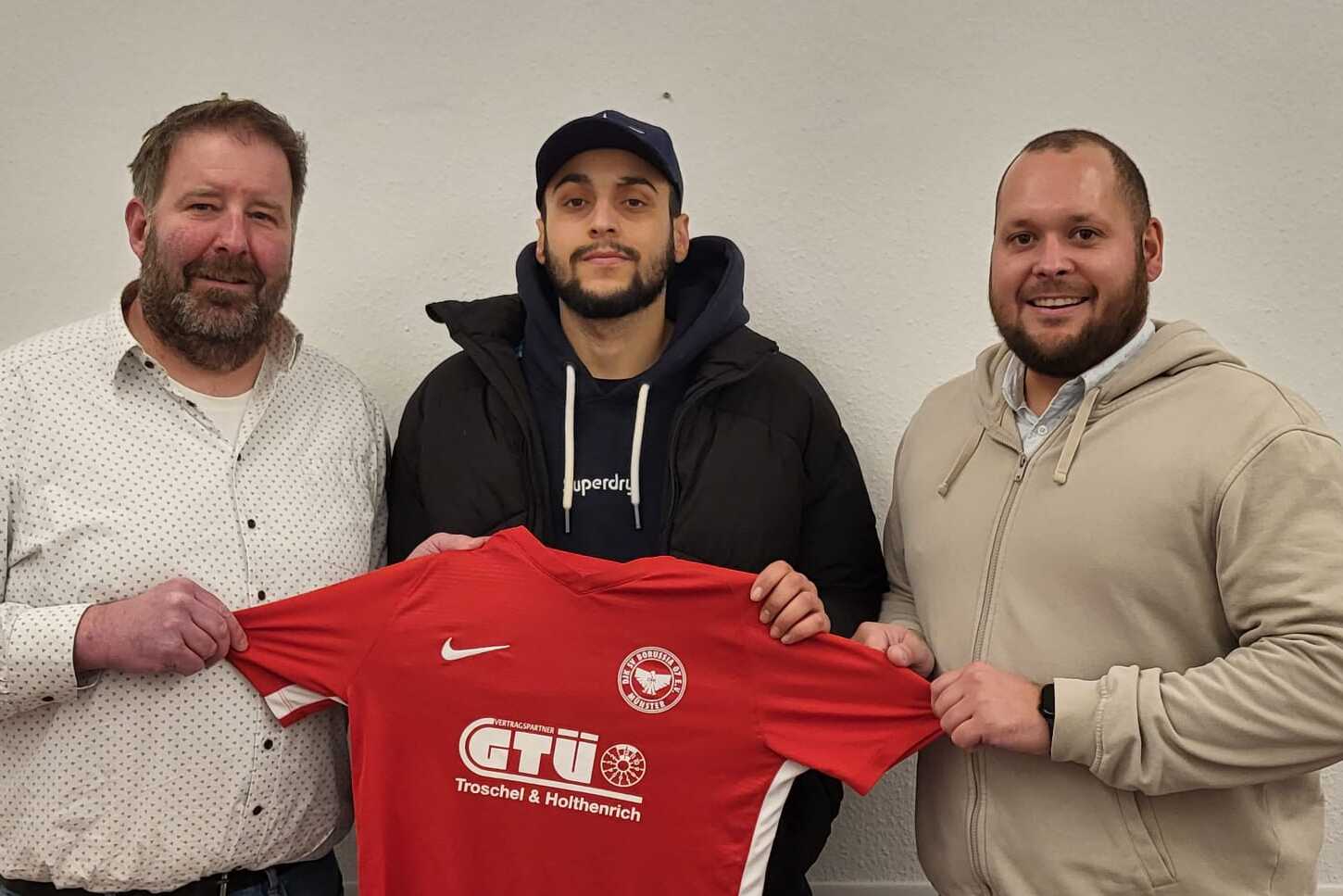 Drei M&auml;nner halten gemeinsam ein rotes Fu&szlig;balltrikot mit Logo, vor einem neutralen Hintergrund.