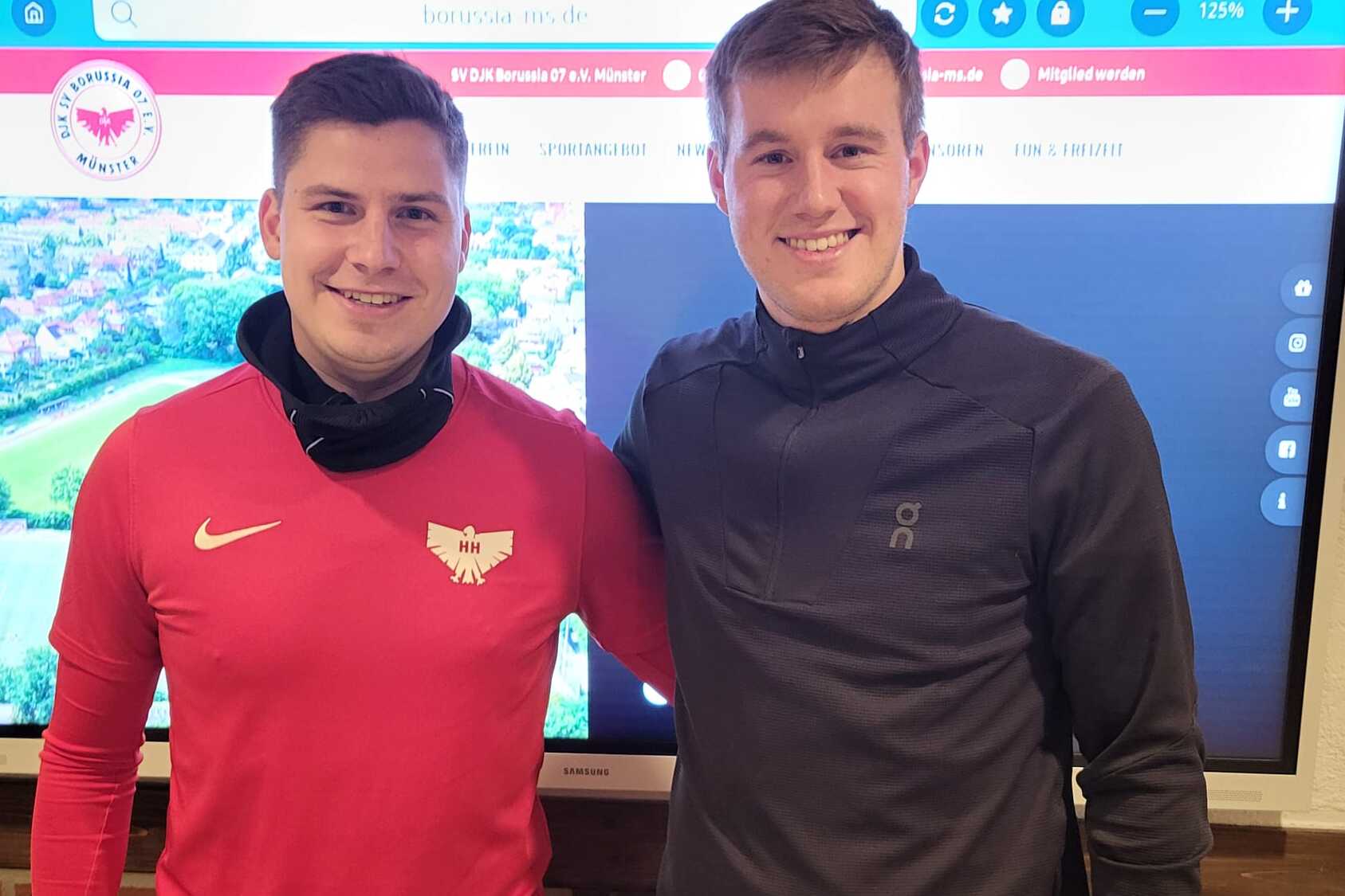 Zwei junge M&auml;nner stehen nebeneinander vor einem Bildschirm mit einer Stadtansicht, einer tr&auml;gt ein rotes Shirt, der andere ein graues.