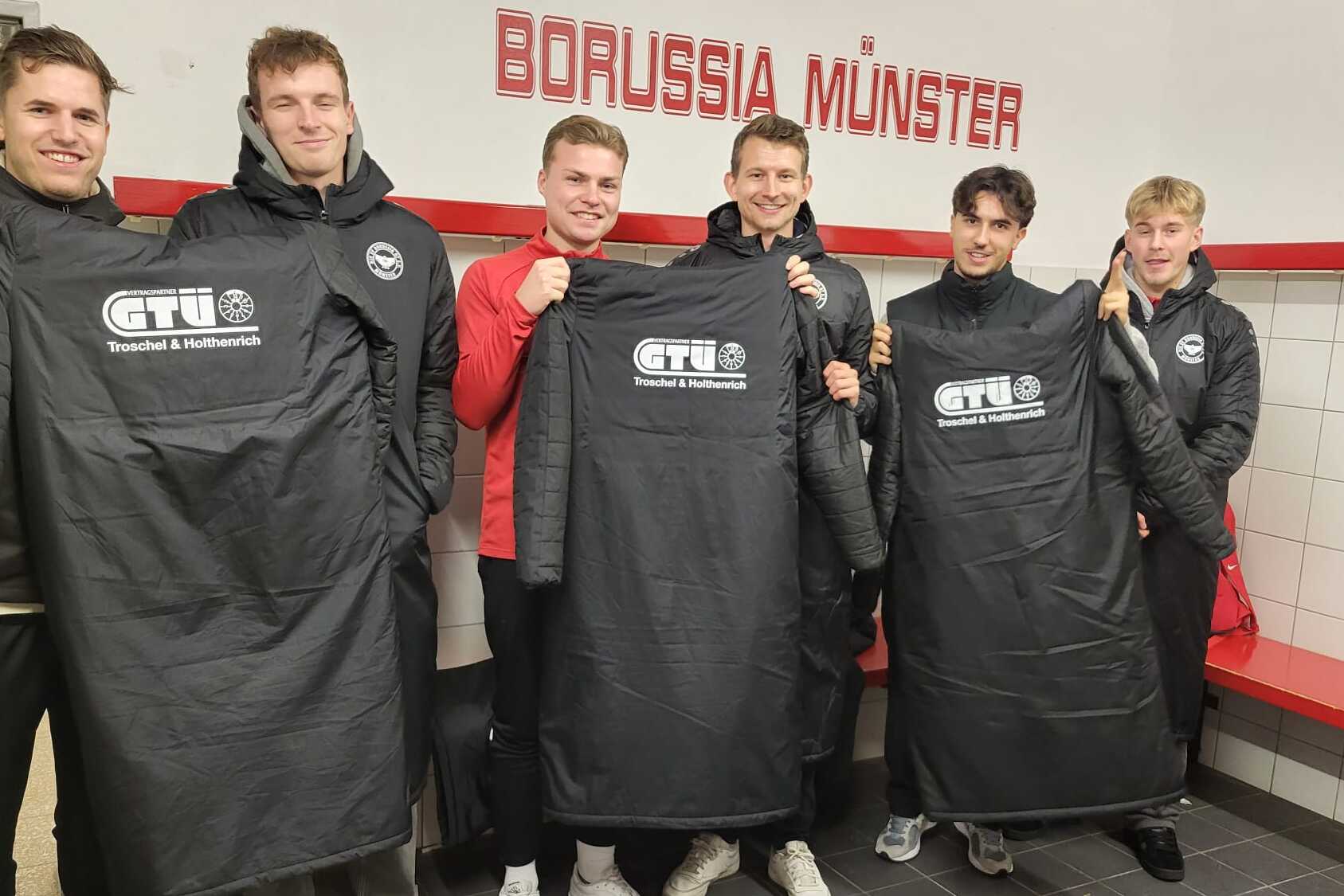 Sechs Sportler in Umkleidung halten schwarze Trikots mit Logo vor dem Schriftzug "BORUSSIA M&Uuml;NSTER" in einem Kabinensaal.