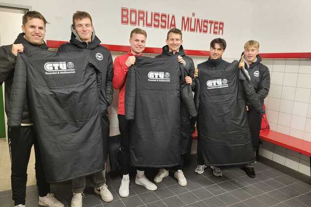 Sechs Sportler in Umkleidung halten schwarze Trikots mit Logo vor dem Schriftzug "BORUSSIA M&Uuml;NSTER" in einem Kabinensaal.