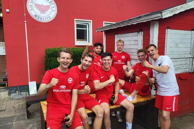 Sechs Fu&szlig;ballspieler in roten Trikots posieren l&auml;chelnd auf einer Bank vor einem roten Vereinsgeb&auml;ude, mit Getr&auml;nken in der Hand.