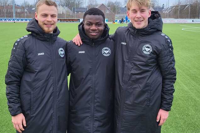 Drei Fu&szlig;ballspieler in schwarzen Trainingsjacken stehen l&auml;chelnd auf einem Rasenplatz.