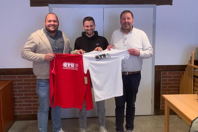 Drei M&auml;nner stehen vor einer geschlossenen T&uuml;r und halten rote und wei&szlig;e T-Shirts mit dem Logo &bdquo;GT&Uuml;&ldquo; in der Hand.