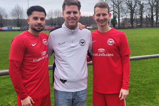 Drei junge M&auml;nner in roten Fu&szlig;balltrikots stehen l&auml;chelnd auf einem Sportplatz mit gr&uuml;ner Wiese im Hintergrund.
